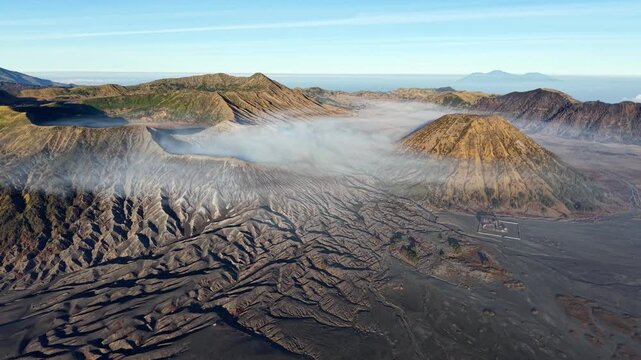 Steam rises from volcanic terrain under clear sky. Rugged, dark earth forms jagged at Bromo