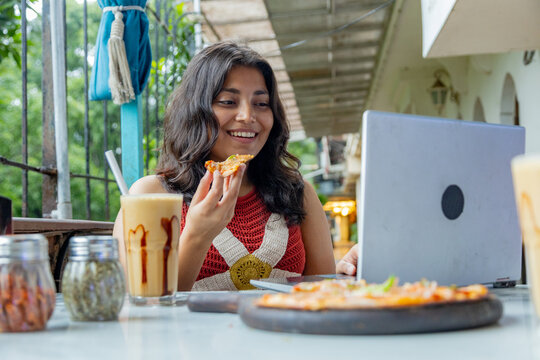 Young indian gen z woman working on laptop while enjoying a snack and a coffee milkshake at a restaurant or cafe, Concept of modern digital nomad lifestyle and remote work flexibility, freelancing