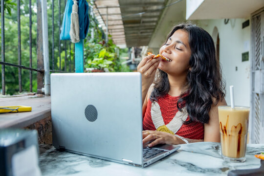 Young indian gen z woman working on laptop while enjoying a snack and a coffee milkshake at a restaurant or cafe, Concept of modern digital nomad lifestyle and remote work flexibility, freelancing