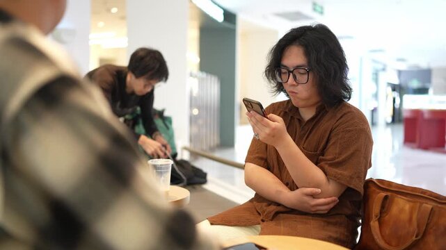 A man in a brown shirt raises his smartphone while another man sits across from him at the table.