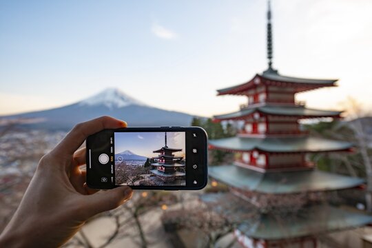 Hand holding smartphone, photographing five-story pagoda of a Shinto shrine, Chureito Pagoda, with views of Fujiyoshida City and Mount Fuji volcano at sunset, Arakura Fuji Sengen Shrine, Arakurayama Sengen Park, Yamanashi Prefecture, Japan