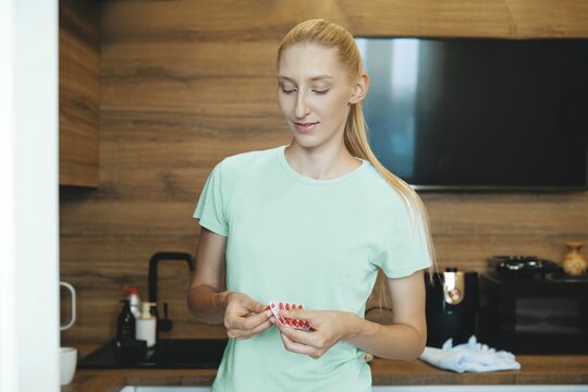 Woman with iron deficiency squeezes pill from package while standing in bright kitchen