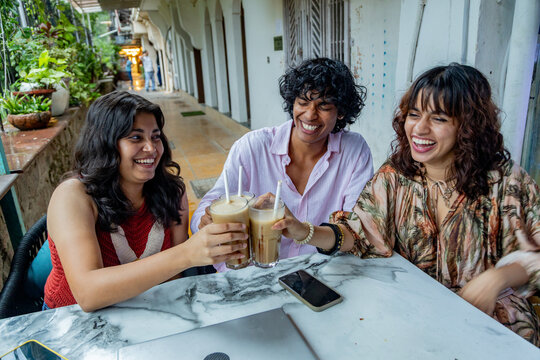 Young indian Gen Z friends enjoying a cheerful moment, clinking glasses of iced coffee at an outdoor cafe, celebrating friendship and social connection during a summer day
