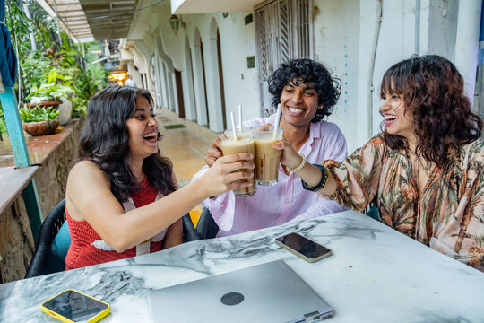 Young indian Gen Z friends enjoying a cheerful moment, clinking glasses of iced coffee at an outdoor cafe, celebrating friendship and social connection during a summer day