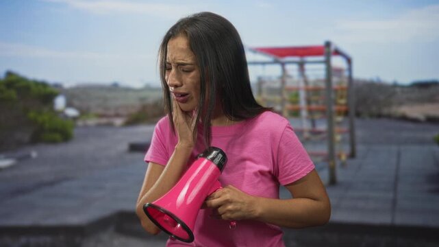 Young hispanic woman holding bright pink megaphone with hand on cheek on building rooftop at dusk; toothache pain.