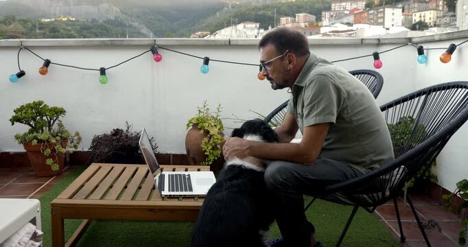 Terrace Serenity: A middle-aged man works on his laptop on a sunny terrace, his dog resting affectionately beside him, creating a scene of relaxed work-life balance with a hint of city view.