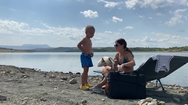 A little boy stands beside his mother as she searches in a bag by the lake. They relax on the beach under a bright cloudy sky. The quiet moment brings a peaceful feeling of warmth and love.