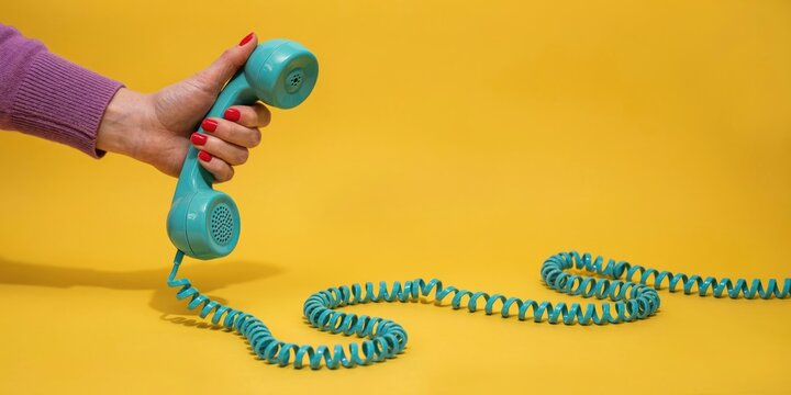 A person's left hand with red nail polish holding a vintage teal telephone handset against a solid yellow background