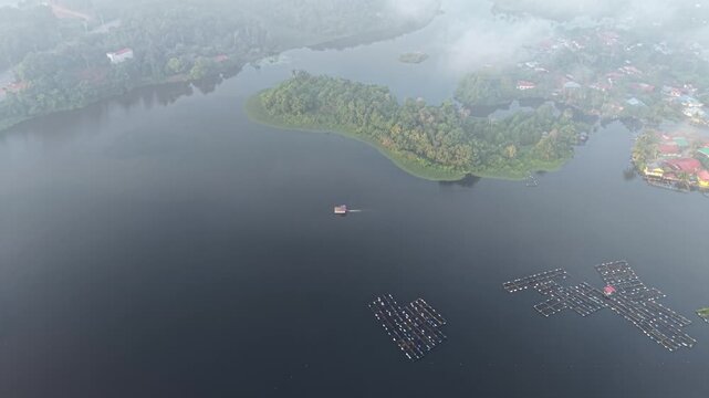A misty aerial view captures a calm lake with floating wooden platforms surrounded by lush green forest and softly enveloped at Tasik Raban
