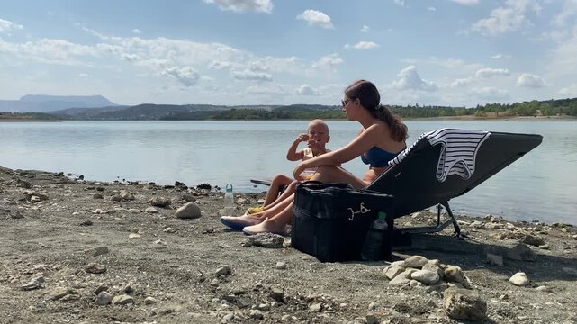 A mother and her young boy sit together on a beach lounger while sharing a snack. They relax by a calm lake under a cloudy sky. The sweet moment brings a warm sense of love and comfort.