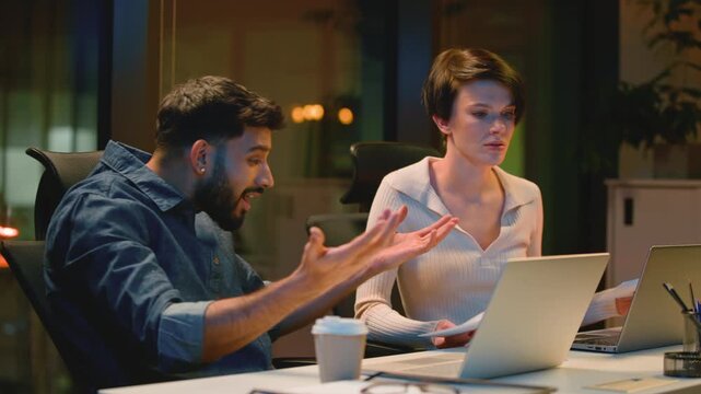 Two multiethnic business colleagues upset with papers documents and laptop at night office. Multiracial coworkers angry Arabian boss man dissatisfied with Caucasian intern woman bad work at evening