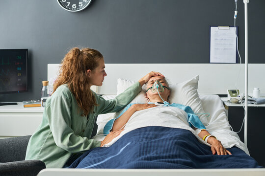 Caucasian young adult woman comforting sick senior woman lying in hospital bed with oxygen mask. Suitable for healthcare campaigns and medical insurance