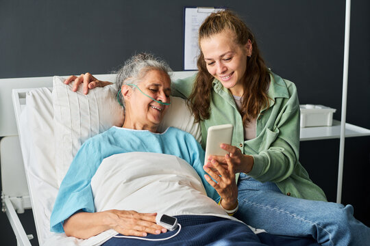 Young Caucasian woman showing smartphone to smiling senior patient in hospital bed. Suitable for healthcare, telemedicine, family support, and medical communication campaigns