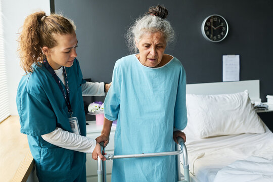 Young adult Caucasian nurse assisting senior Caucasian female patient walking with walker frame in clinic room. Concept suitable for geriatric healthcare advertising