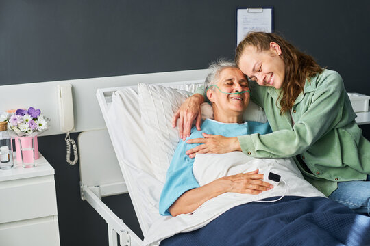 Young adult Caucasian woman hugging smiling senior Caucasian female patient in hospital bed. Image represents family support, recovery care, suitable for medical insurance advertising