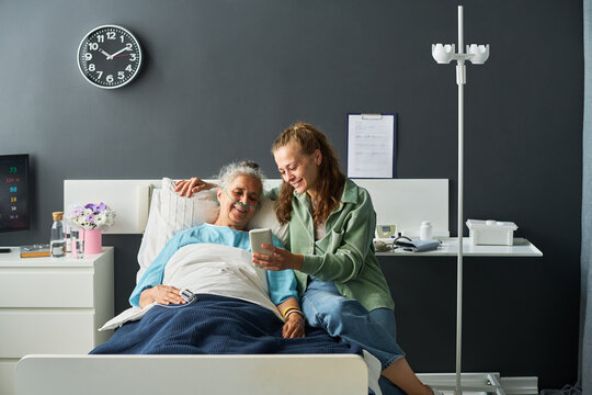 Senior woman resting in hospital bed looking at smartphone with young adult Caucasian visitor. Suitable for telemedicine campaigns and senior healthcare marketing