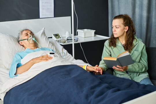 Caucasian young adult woman holding hands with sick senior Caucasian woman wearing oxygen mask in hospital ward. Ideal for healthcare campaigns and insurance media