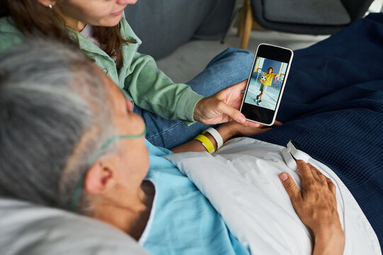 Young adult woman showing smartphone screen with child photo to senior Caucasian hospital patient in bed. Concept represents family support, suitable for healthcare advertising