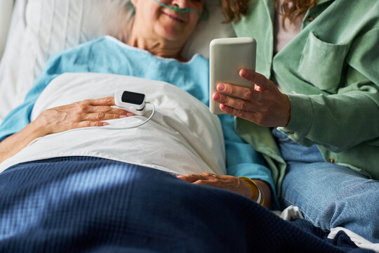 Senior Caucasian woman lying in hospital bed while adult daughter shows smartphone screen during family visit. Image useful for digital healthcare app and patient communication campaigns