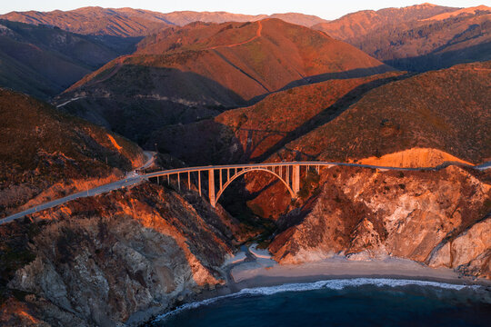 Aerial view captures Bixby Creek Bridge along the Pacific Coast Highway in Big Sur, California, spanning a rocky coastal canyon under warm sunset light with waves meeting the sandy beach below.