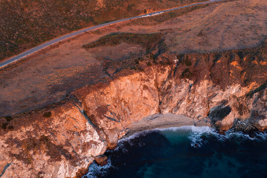 Aerial perspective displays steep rugged cliffs, an isolated sandy cove, and the Pacific Coast Highway bathed in warm sunset light in Big Sur, California.