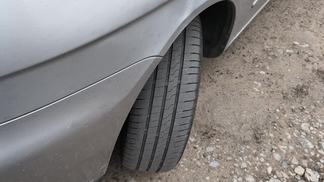 Close-up shot of a car tire resting on a dirt and gravel road. The detailed view highlights the rubber tread pattern and the edge of a grey vehicle bumper. Great for auto maintenance concepts.