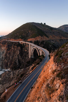 Bixby Creek Bridge curves along Highway 1 on the rugged Pacific coast in Big Sur, California, with distant tourists standing by the roadside at sunset as warm light illuminates the cliffside.
