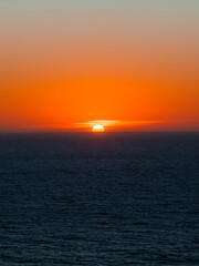 Obraz premium Sun sets directly into the Pacific Ocean horizon viewed from the Big Sur coast in California, illuminating the evening sky with warm orange tones above gentle waves.
