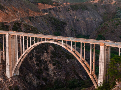 Aerial shot of Bixby Creek Bridge spanning a steep canyon along the Pacific Coast Highway in Big Sur, California, illuminated by warm evening sunlight.