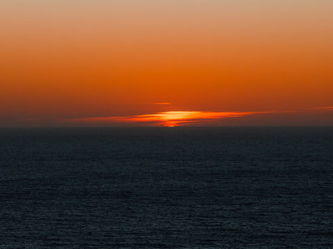 Bright orange sunset lights up the evening sky over dark Pacific Ocean waters off the coast of Big Sur in California. The glowing sun dips below a line of clouds along the horizon.
