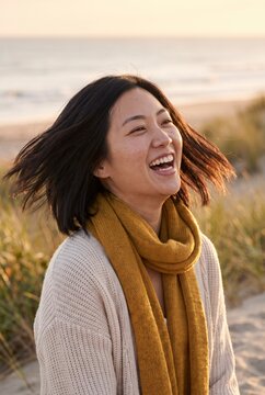 Joyful Asian woman laughing with windswept hair on golden beach dunes at sunset