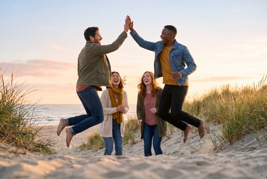 Diverse friends jumping for a joyful high five together on beach dunes at sunset