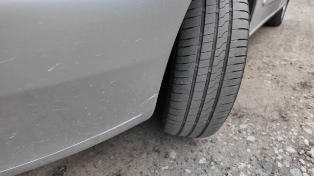 Close-up shot of a car tire resting on a dirt and gravel road. The detailed view highlights the rubber tread pattern and the edge of a grey vehicle bumper. Great for auto maintenance concepts.