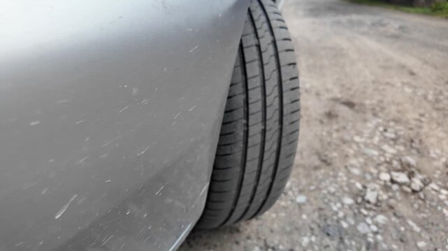 Close-up shot of a car tire resting on a dirt and gravel road. The detailed view highlights the rubber tread pattern and the edge of a grey vehicle bumper. Great for auto maintenance concepts.