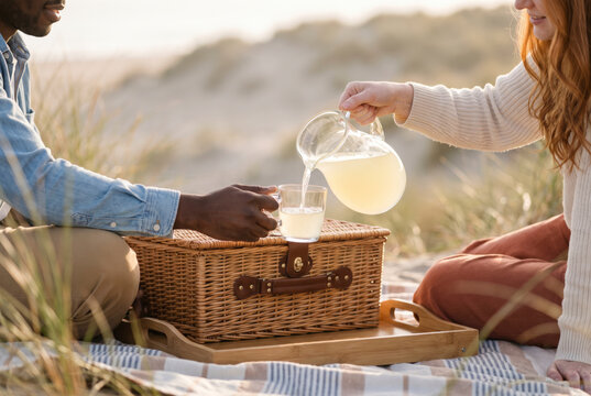 Hands pouring lemonade into a glass on wicker picnic basket at sunset beach