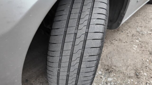 Close-up shot of a car tire resting on a dirt and gravel road. The detailed view highlights the rubber tread pattern and the edge of a grey vehicle bumper. Great for auto maintenance concepts.
