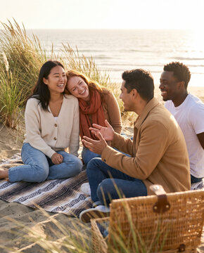 Diverse friends chatting together around picnic basket on beach dunes at sunset