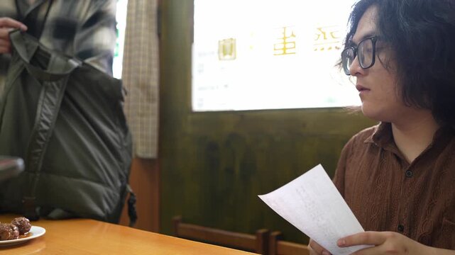A seated man handles a folded paper item at a restaurant table while another person stands nearby.