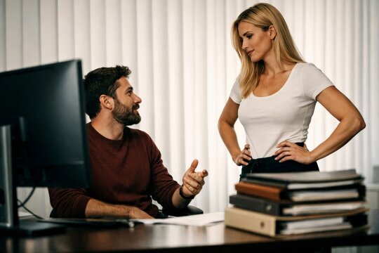Office colleagues discussing work at desk with computer and folders, professional conversation in modern workplace.