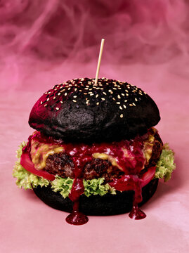 Black burger with dripping sauce and sesame seed bun on pink background