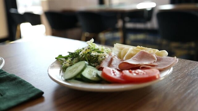  Detailed view of a delicious morning meal or buffet plate served in a cafe, featuring sliced ham, fresh cucumbers, cheese, and salad greens on a wooden surface.