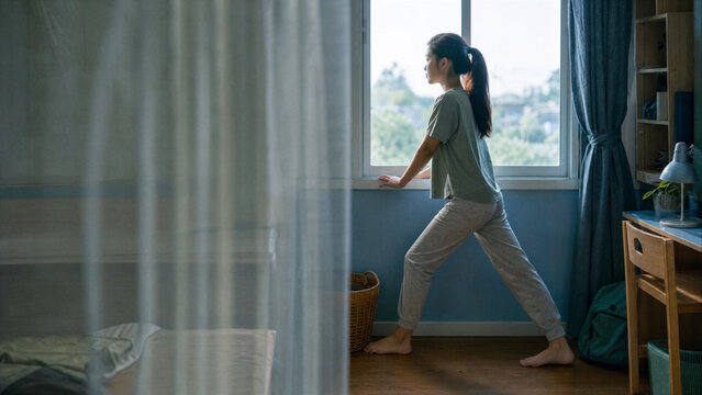Asian woman stretching her body near open window in cozy bedroom. Young girl in pajamas doing morning exercise at home in soft daylight.