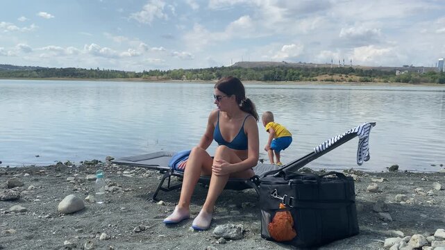 A mother sits on a lounge chair by a calm lake while her toddler stands near the water. They share a peaceful and relaxing moment, feeling calm and content in nature.