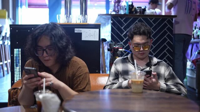 Two men sit at a bar or restaurant table with drinks while both look down at smartphones.