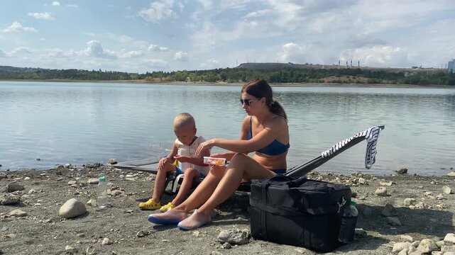 A loving mother and her little son share a small snack while sitting on a lounger. They relax together on a rocky shore beside a calm lake. Their gentle bonding moment feels warm and peaceful.