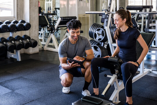 Personal trainer coaches a woman on a gym machine, reviewing her workout on a tablet among weights in a modern fitness studio.