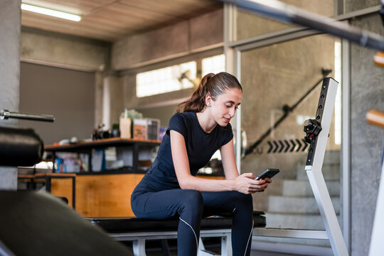 Woman in athletic wear sits on a gym bench checking her smartphone during a workout break, surrounded by fitness equipment in a modern training space.
