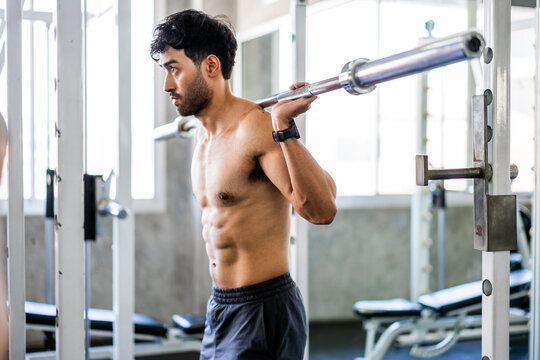 Shirtless man preparing to back squat with a barbell in a gym rack, focused and ready to lift