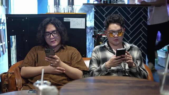 Two men sit side by side at a bar or restaurant table and look down at their smartphones.