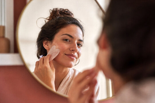 Young woman massages her cheek with a gua sha tool, reflected in a round mirror. She has clear skin and a radiant smile, practicing self-care.